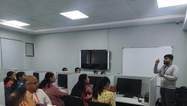 Man standing at right speaks to seated participants at computer cubicles in a lab, with a whiteboard and wall screens behind him.