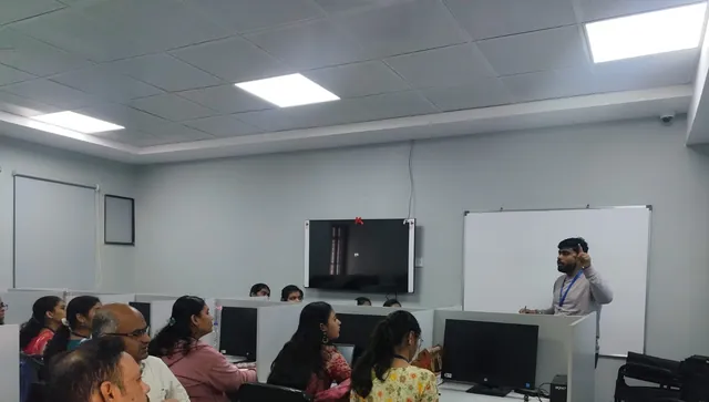 A man stands by a whiteboard and speaks to a group of adults seated at computer workstations in a lab.