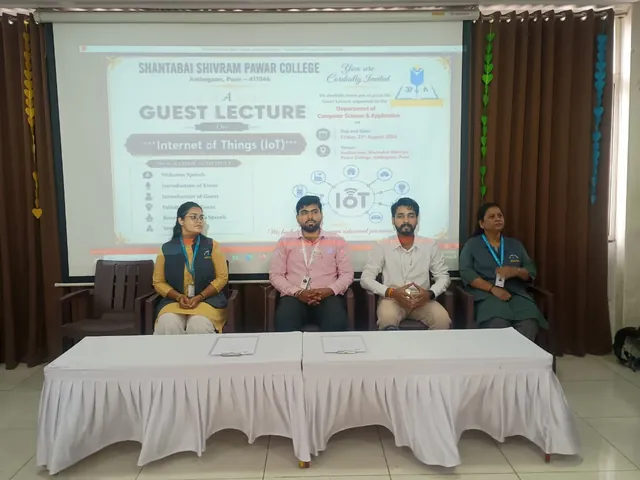 Four people seated behind a white-draped table in front of a projected slide announcing a guest lecture on the Internet of Things (IoT).