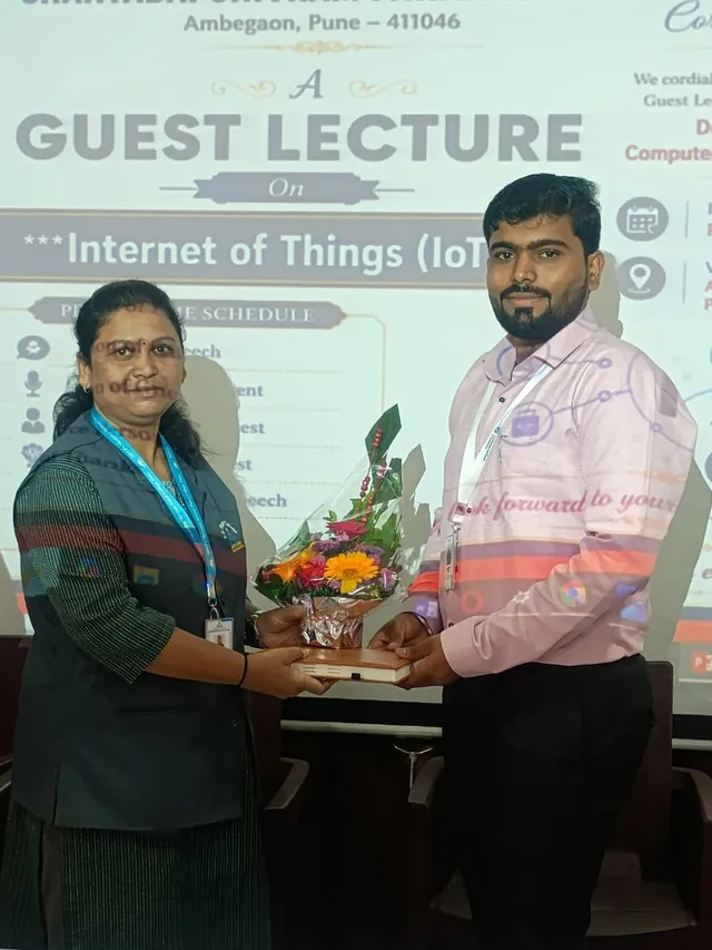 A woman and a man in a pink shirt hold a flower bouquet and a book in front of a projected Guest Lecture on Internet of Things slide.