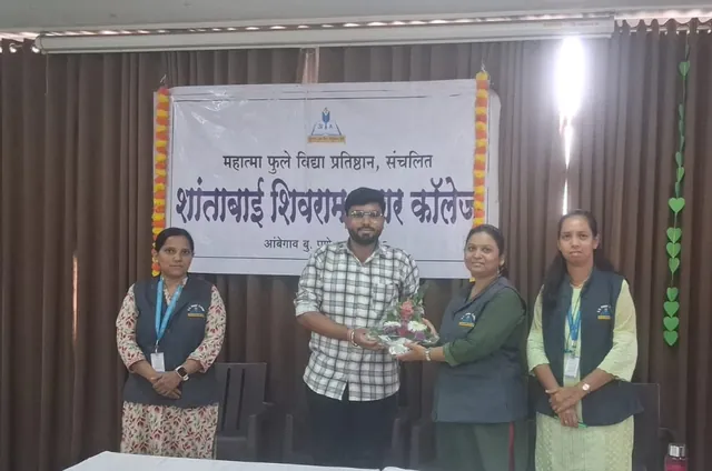 Man in a checked shirt receives a flower bouquet from a woman, with two women beside them, in front of a Marathi college banner.