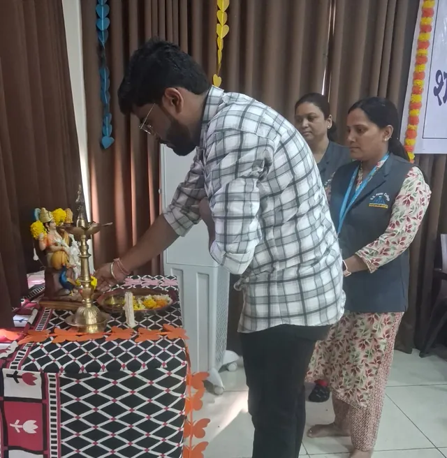 Man lighting a brass lamp beside a Saraswati idol while two women in grey vests stand behind him.
