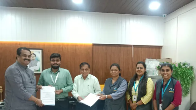 Wider view of six people behind a desk in an office, two pairs each holding a document between them.